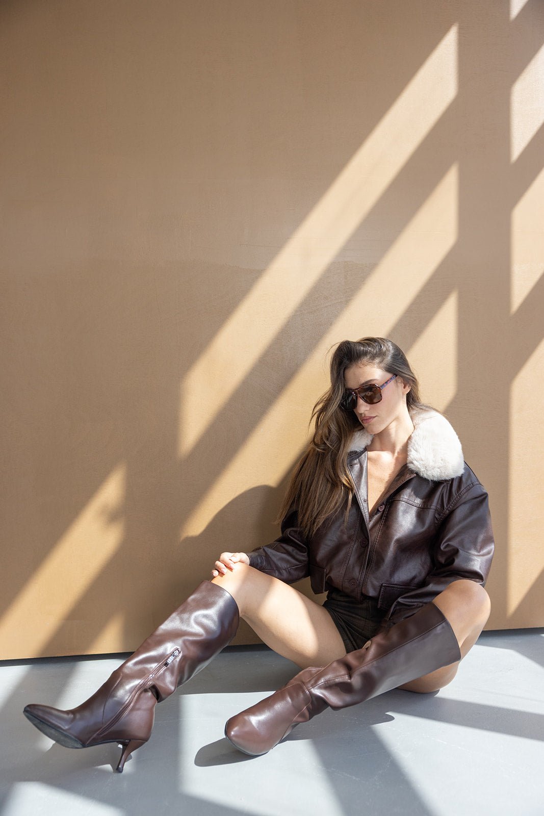 דוגמנית עם מעיל עור חום עם צווארון פרווה, מגפיים חומים ומשקפי שמש, יושבת בשמש