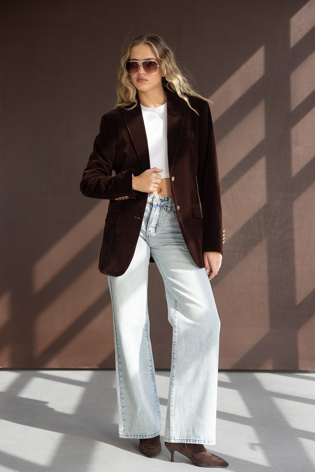 אישה לובשת בלייזר חום, ג'ינס בגזרה רחבה, חולצה לבנה קצרה ומשקפי שמש אופנתיות