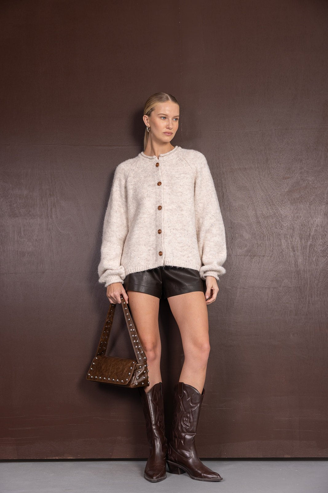 אישה עם סוודר קרמי מכופתר, מכנסי עור קצרים, מגפיים חומים ותיק תואם, אופנה לנשים