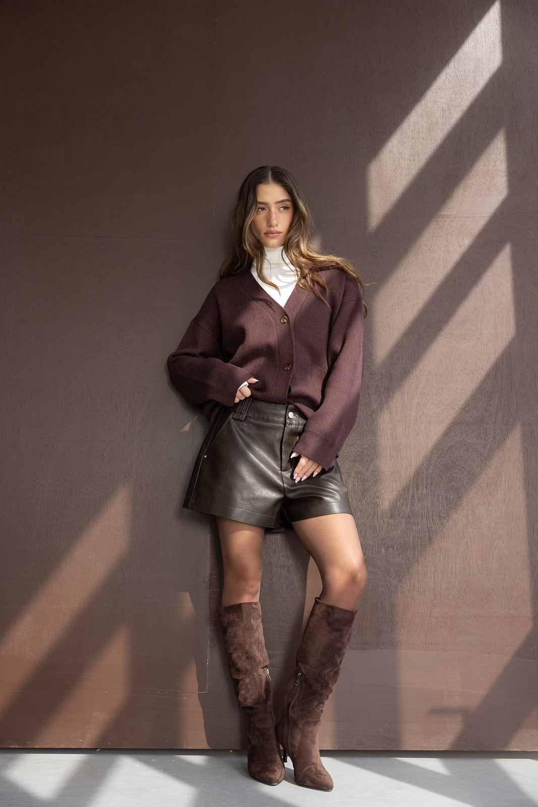 אישה צעירה לובשת סוודר בורדו, חצאית עור חומה, מגפיים חומים גבוהים, אופנה חורפית מודרנית