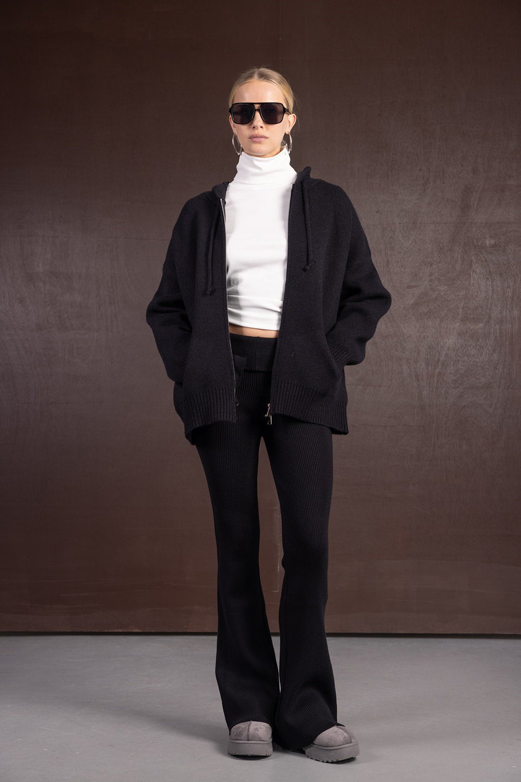 אישה עם גופיית גולף לבנה, סוודר שחור, מכנסיים שחורים רחבים ונעלי בית אפורות, לוק חורפי אופנתי.