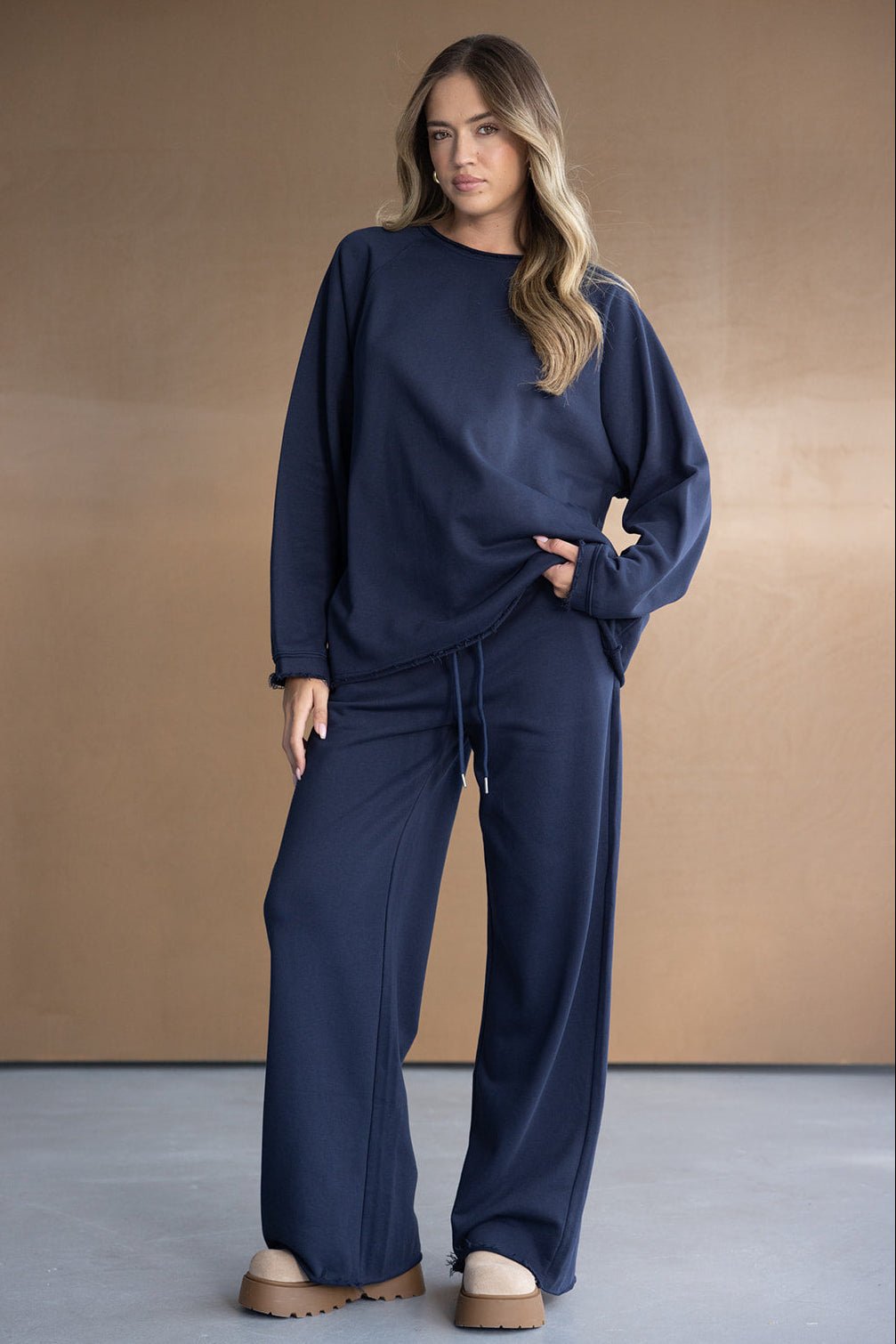 אישה לובשת סט טרנינג כחול כהה, חולצה ארוכה ומכנסיים רחבים, עומדת על רקע ניטרלי