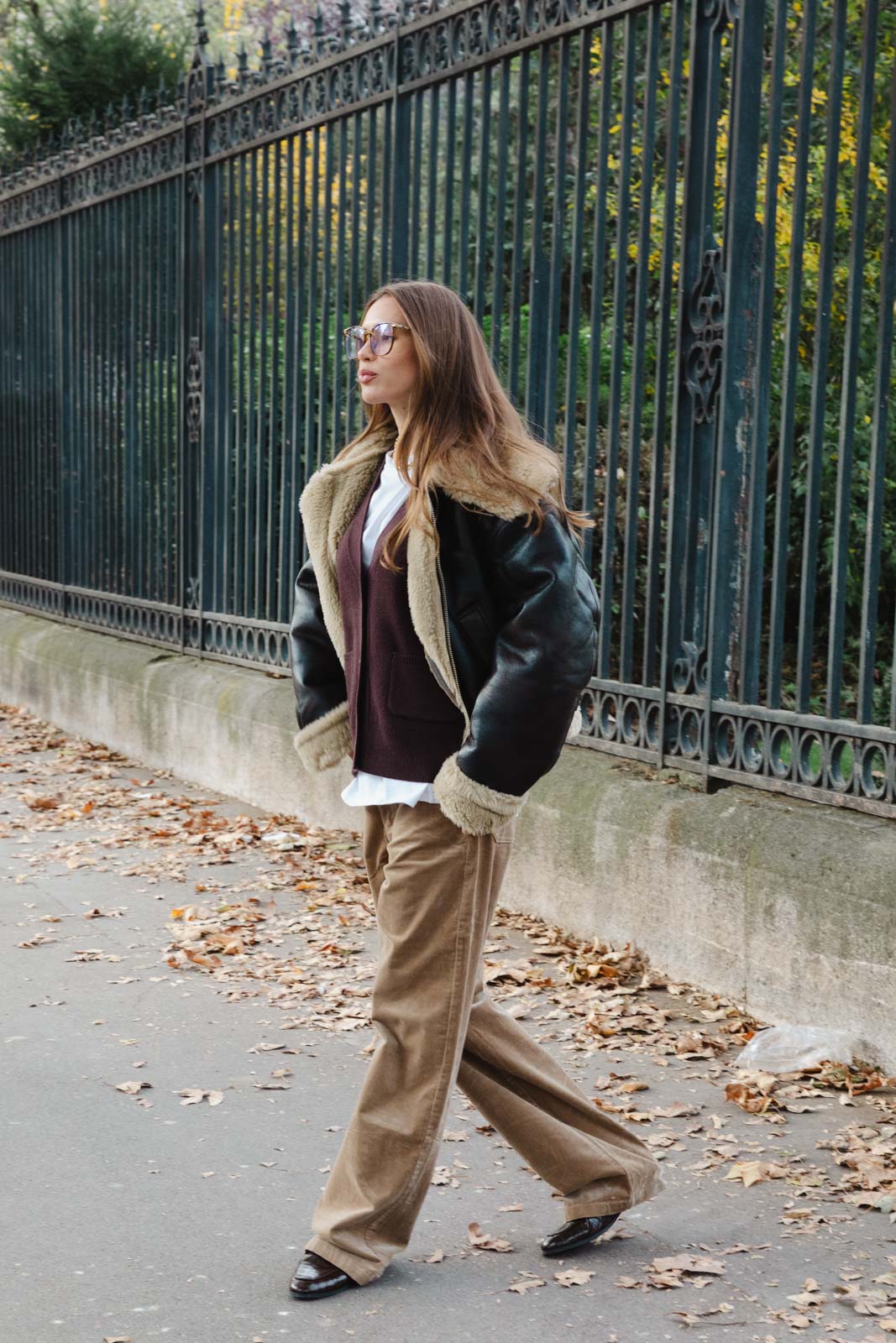 אישה צעירה לובשת מעיל דמוי פרווה, וסט חום ומכנסי קורדרוי אופנתיים, צועדת ברחוב עירוני בסתיו