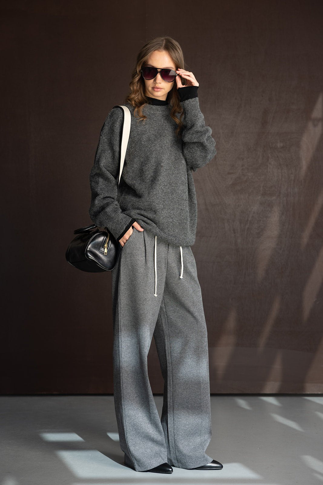 אישה עם סווטשרט אפור ומכנסיים רחבים, משקפי שמש ותיק צד, אופנת חורף לנשים