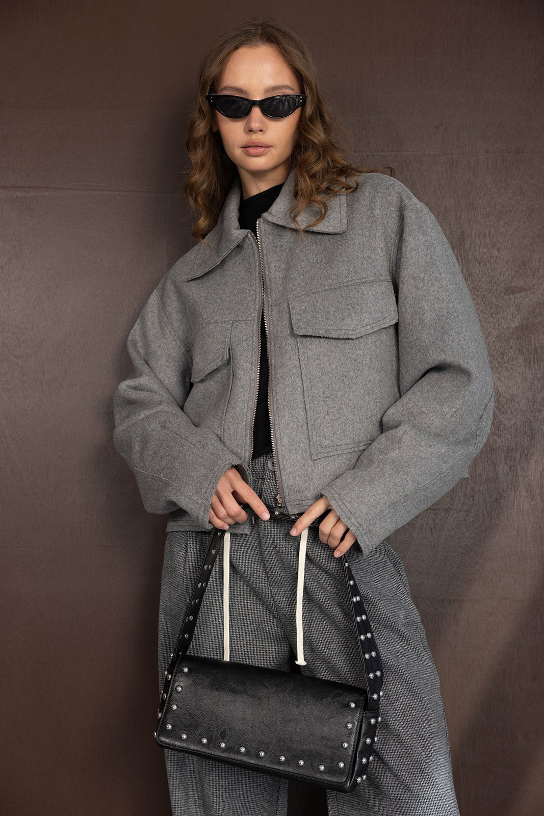 אישה צעירה עם ז'קט אפור אופנתי, מחזיקה תיק שחור עם ניטים, משקפי שמש כהים, לוק חורפי קז'ואל לנשים