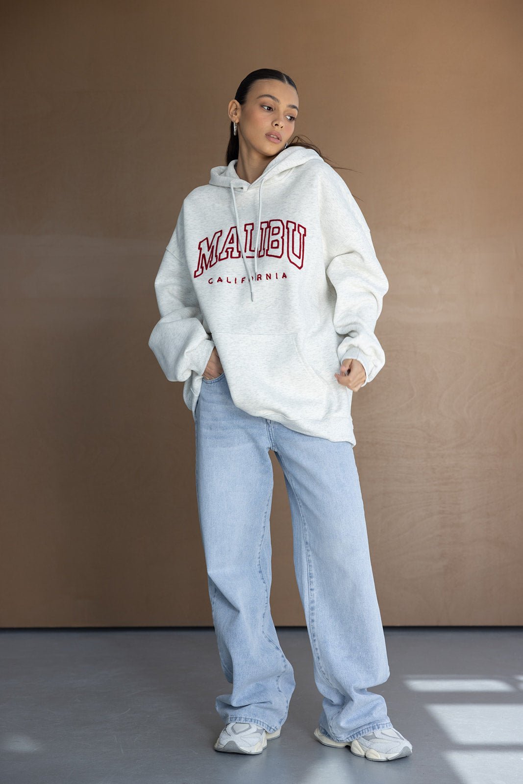 דוגמנית לובשת קפוצ'ון אפור עם כיתוב MALIBU ומכנסי ג'ינס בגזרה רחבה, אופנת רחוב לנשים