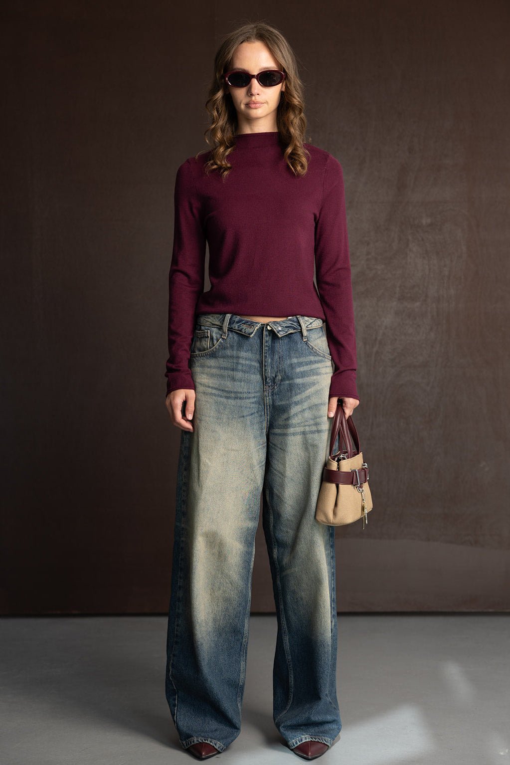 אישה צעירה עם חולצה סגולה ארוכת שרוולים, ג'ינס רחב גבוהה ומשקפי שמש, מחזיקה תיק אופנתי