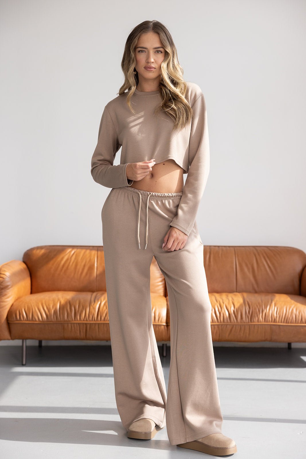 אישה לובשת סט ביגוד ביתי אופנתי בצבע בז', חולצה קצרה ומכנסיים רחבים, ליד ספת עור