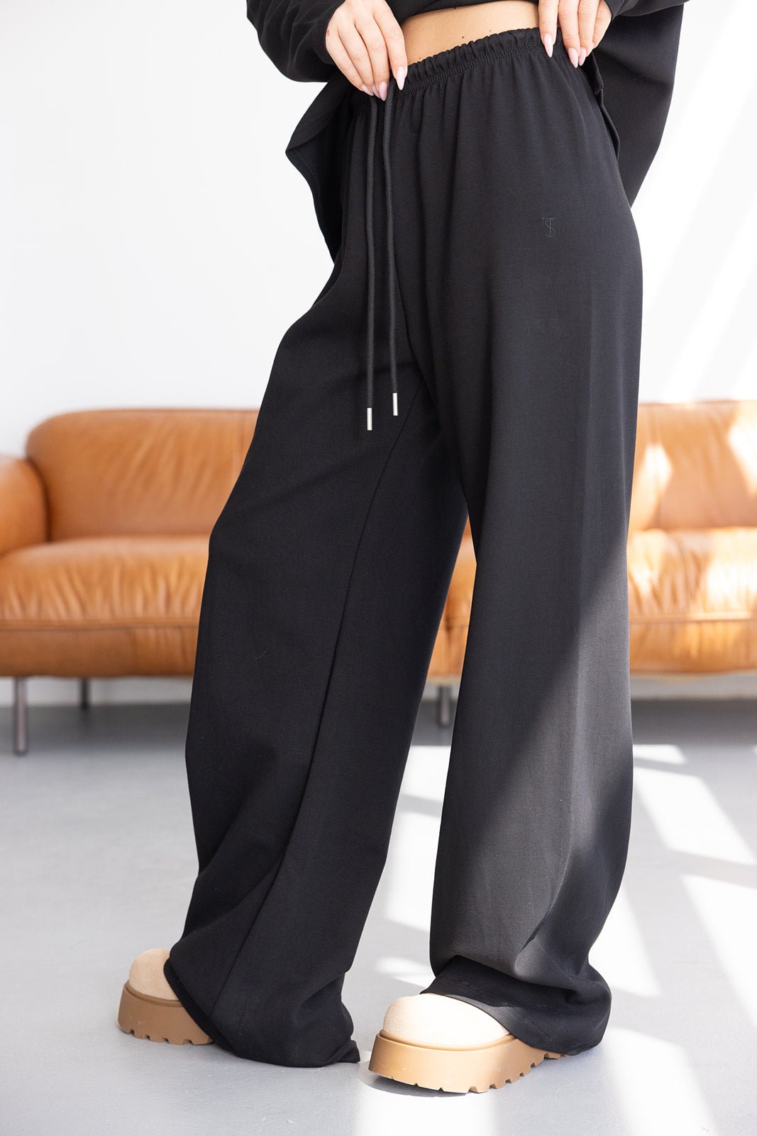מכנסי נשים שחורים רחבים עם גומי ושרוך, לוק יומיומי אופנתי, נעליים טרנדיות ברקע סלון מודרני
