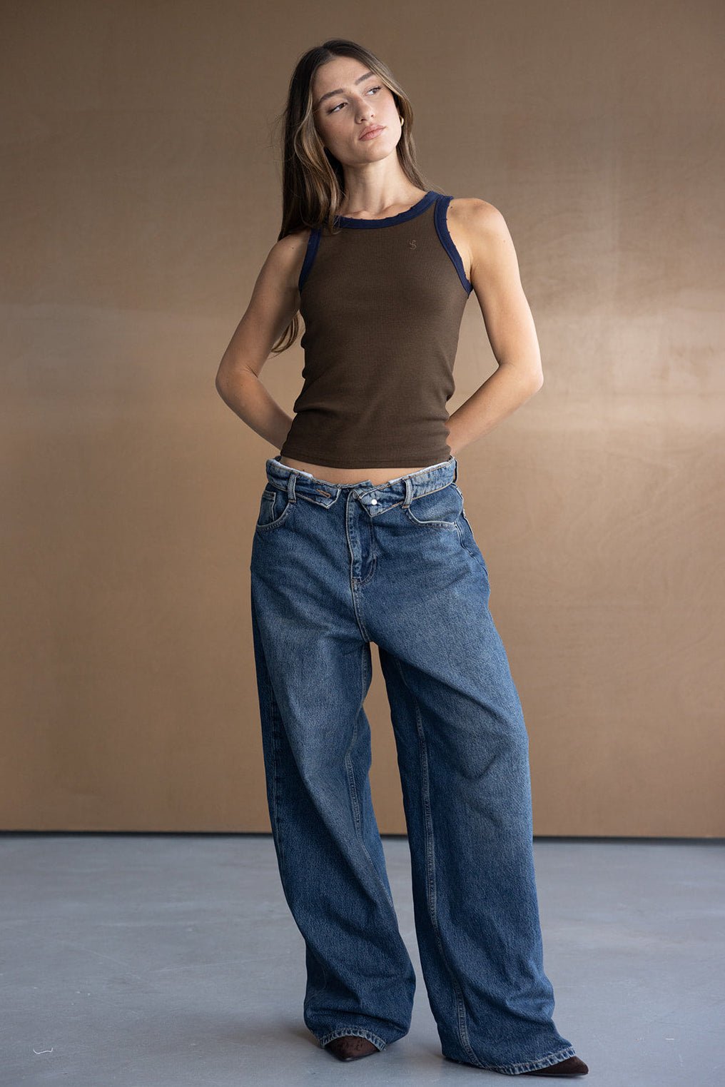 דוגמנית עם גופיית סריג חומה ומכנסי ג'ינס רחבים, אופנה לנשים צעירות, רקע חום