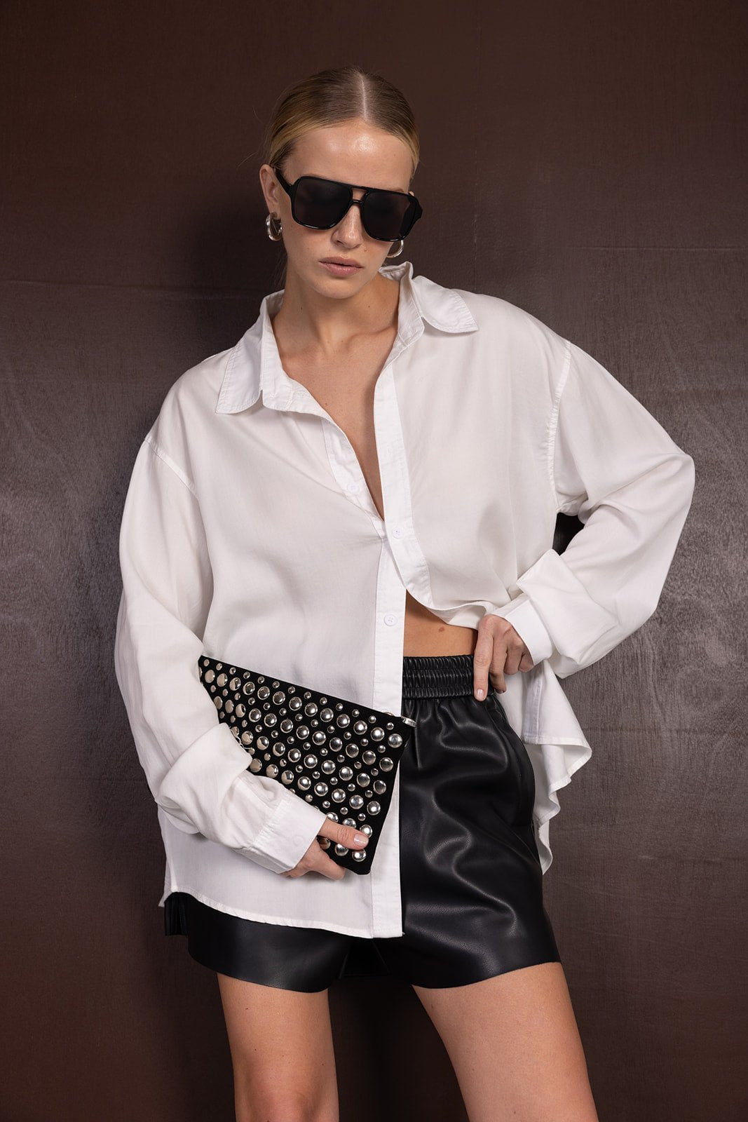 אישה עם חולצת כפתורים לבנה, מכנסי דמוי עור שחורים, משקפי שמש ותיק קלאץ' מעוצב עם ניטים, אופנה לנשים