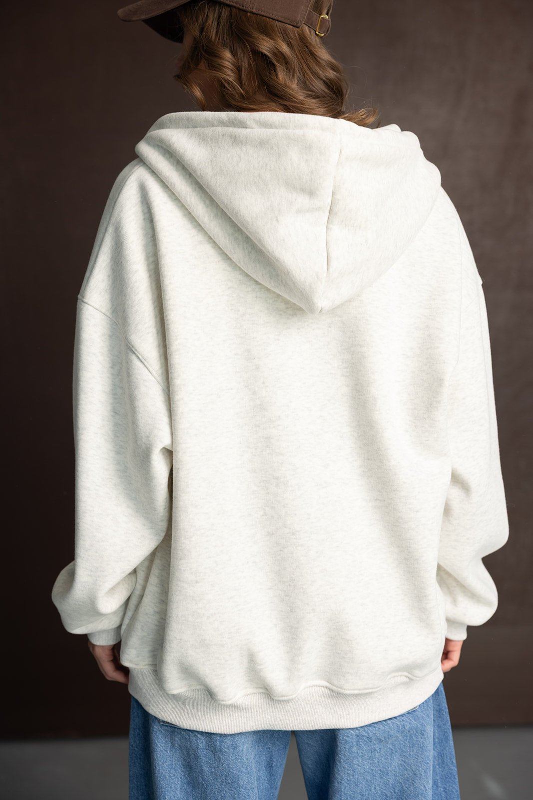 אישה עם סווטשירט קפוצ׳ון אפור בהיר בגזרה רחבה, ג׳ינס כחול, כובע חום, מצולם מאחור