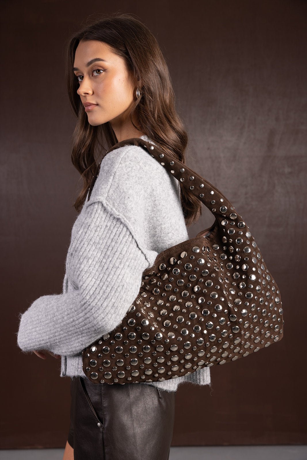 דוגמנית צעירה לובשת סוודר אפור עם חצאית עור ושואלת תיק חום עם ניטים, אופנת נשים עדכנית