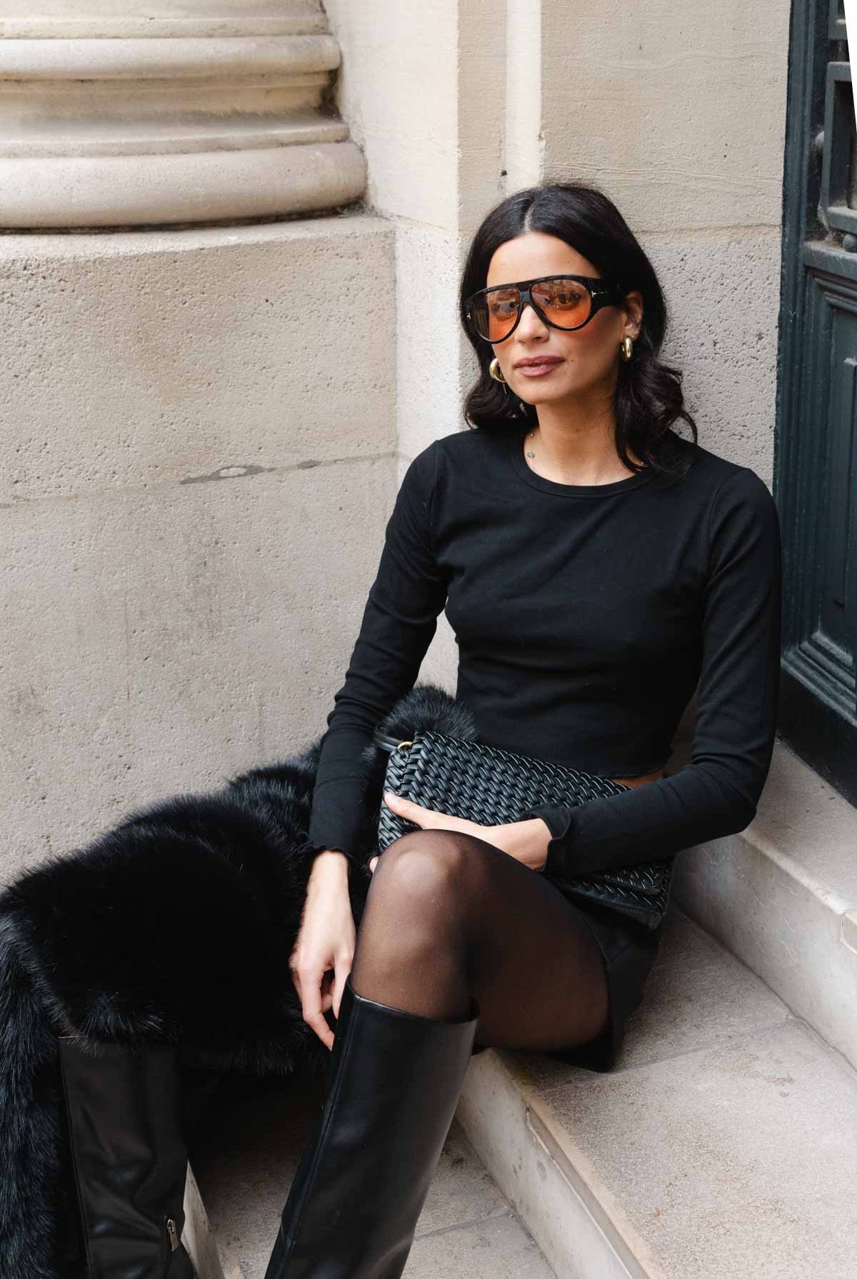 אישה יושבת בלבוש אופנתי, חולצה שחורה, חצאית קצרה, מגפיים גבוהים ותיק ערב, לוק אורבני לנשים