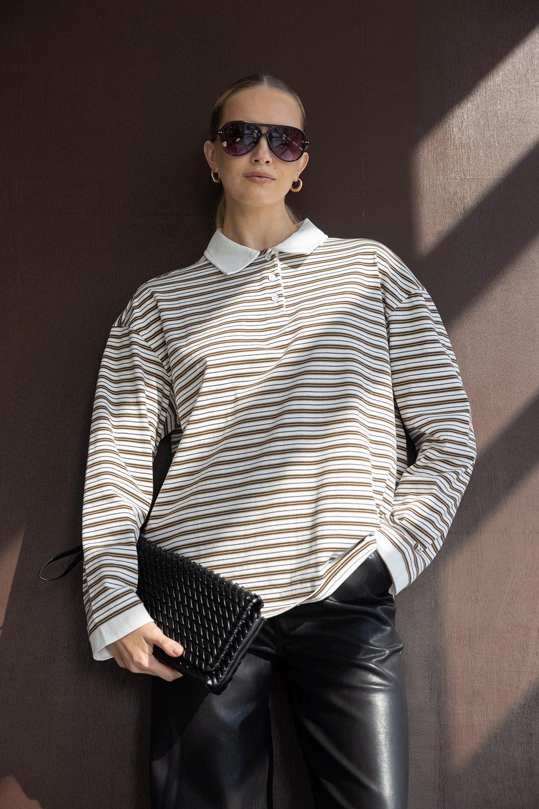 אישה עם חולצת פסים ארוכה, מכנסי דמוי עור שחור, משקפי שמש ותיק קלאץ' אופנתי