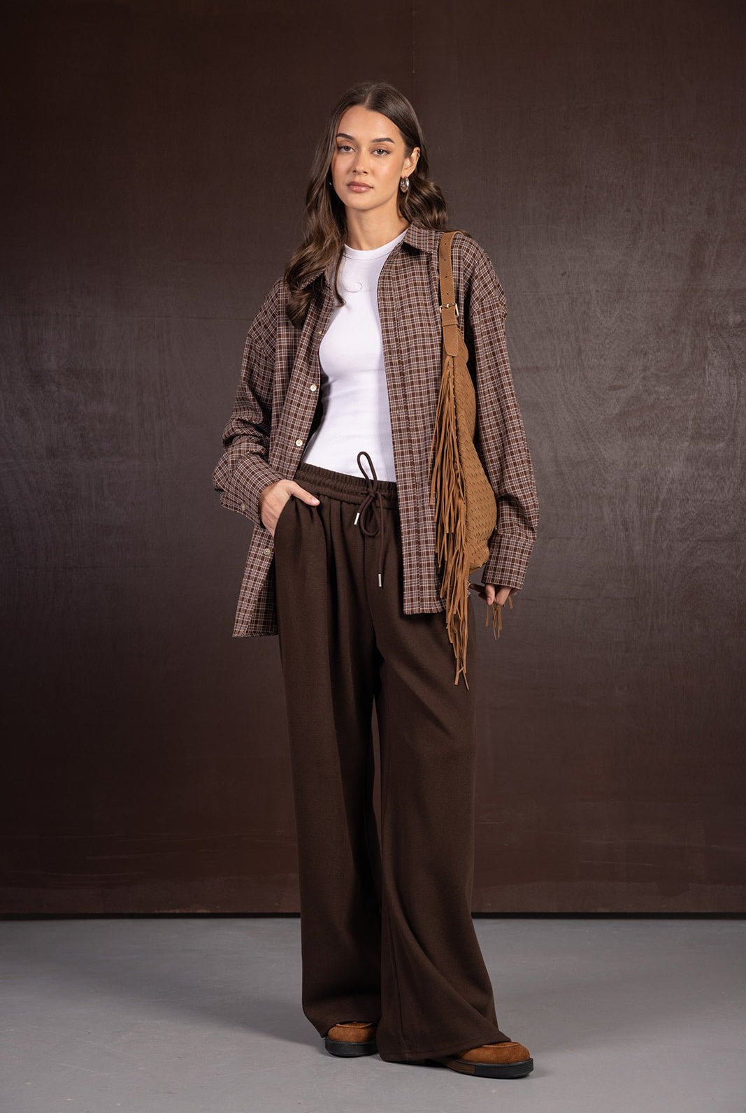 אישה עם חולצה לבנה, חולצת פלנל משובצת חומה, מכנסיים רחבים חומים, תיק צד עם פרנזים ונעלי מוקסין, לוק אופנתי סתיו חורף