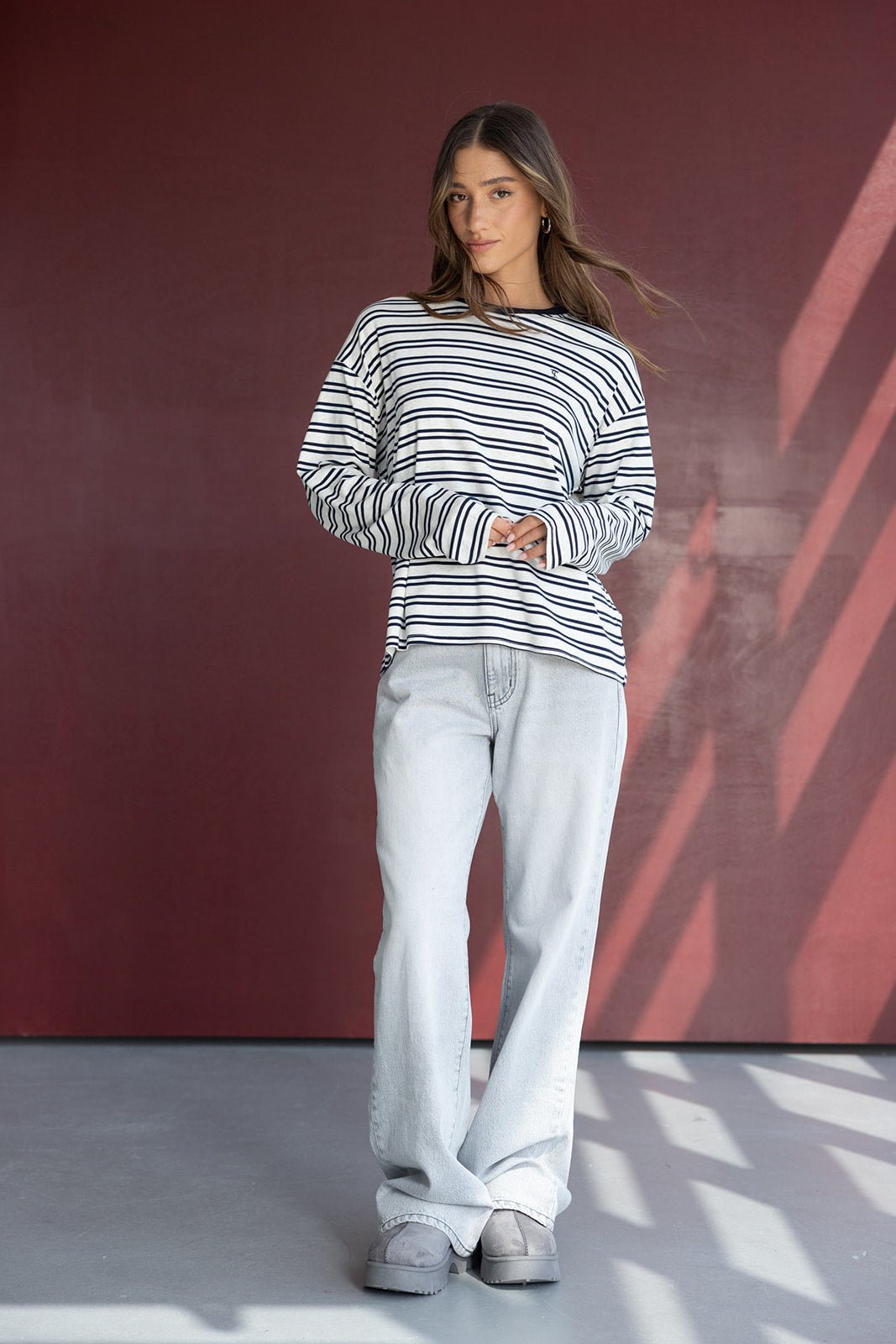 דוגמנית לובשת סווטשירט פסים שחור-לבן רחב עם מכנסי ג'ינס רחבים ונעלי בית אפורות, רקע בורדו.