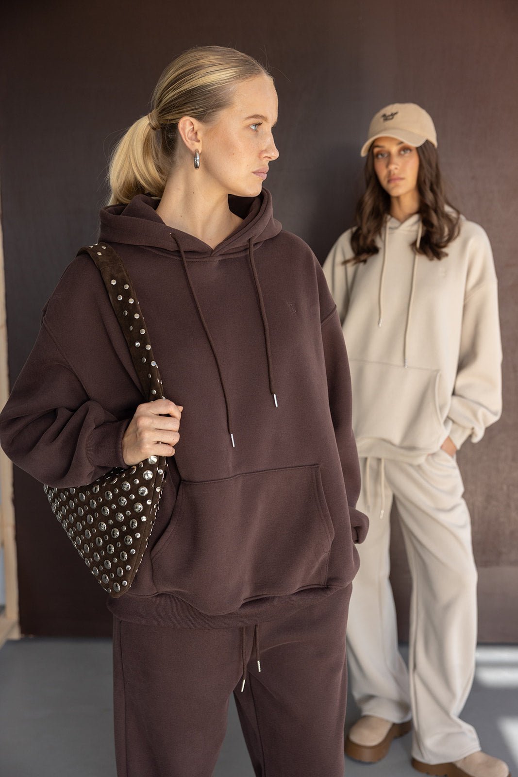 שתי נשים עם הודי ומכנסיים רחבים, אחת בחליפת טרנינג חומה עם תיק ניטים, השנייה בלבוש בז' וכובע מצחייה, אופנת רחוב לנשים