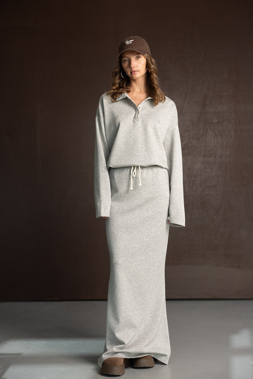 דוגמנית עם שמלת סווטשירט אפורה ארוכה, כובע קפטן חום ונעלי פלטפורמה, צילומי אופנה ברקע חום