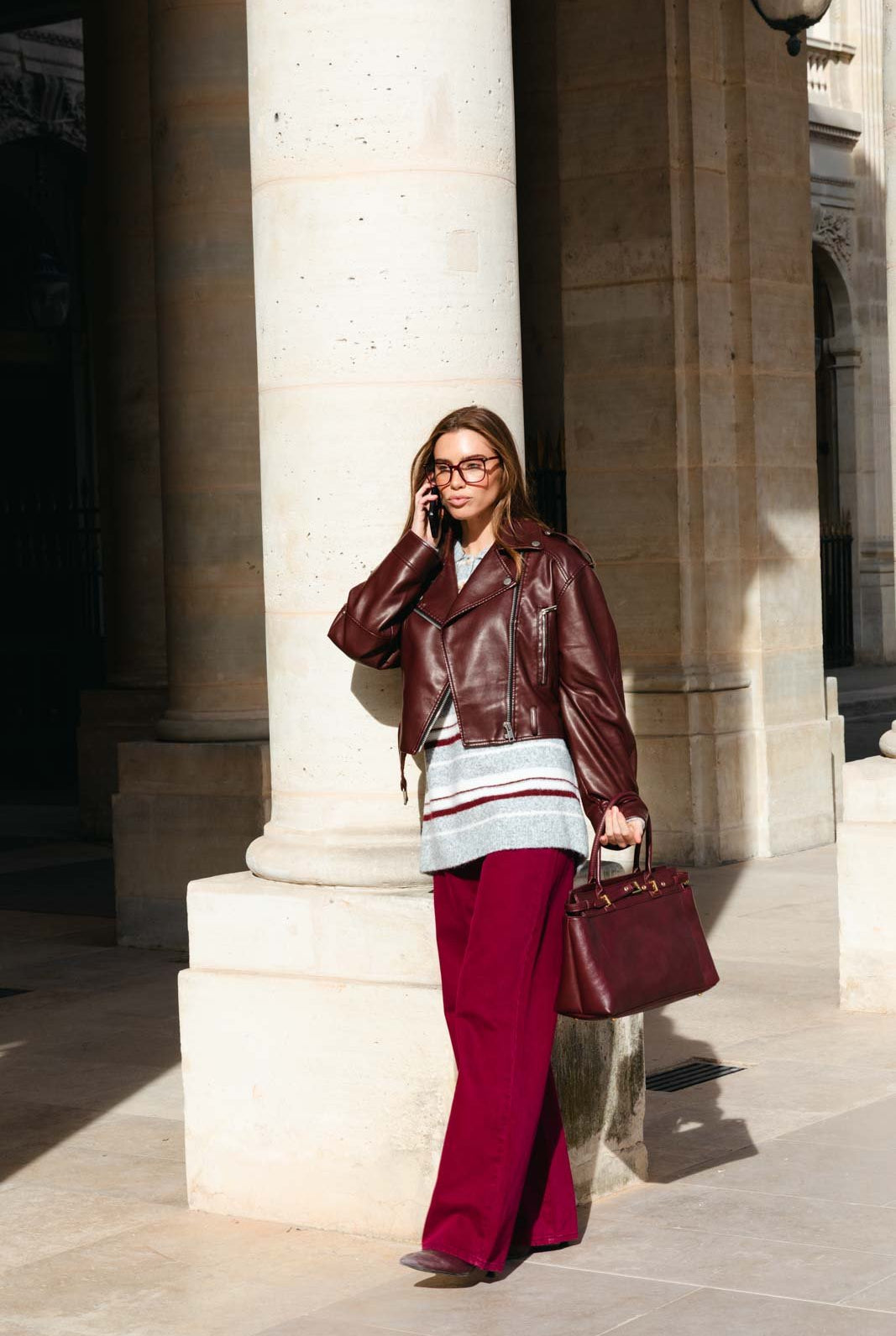 אישה בבגדי אופנה, מעיל עור חום, מכנסיים רחבים ובגד סריג, מחזיקה תיק ומתהלכת ברחוב עירוני