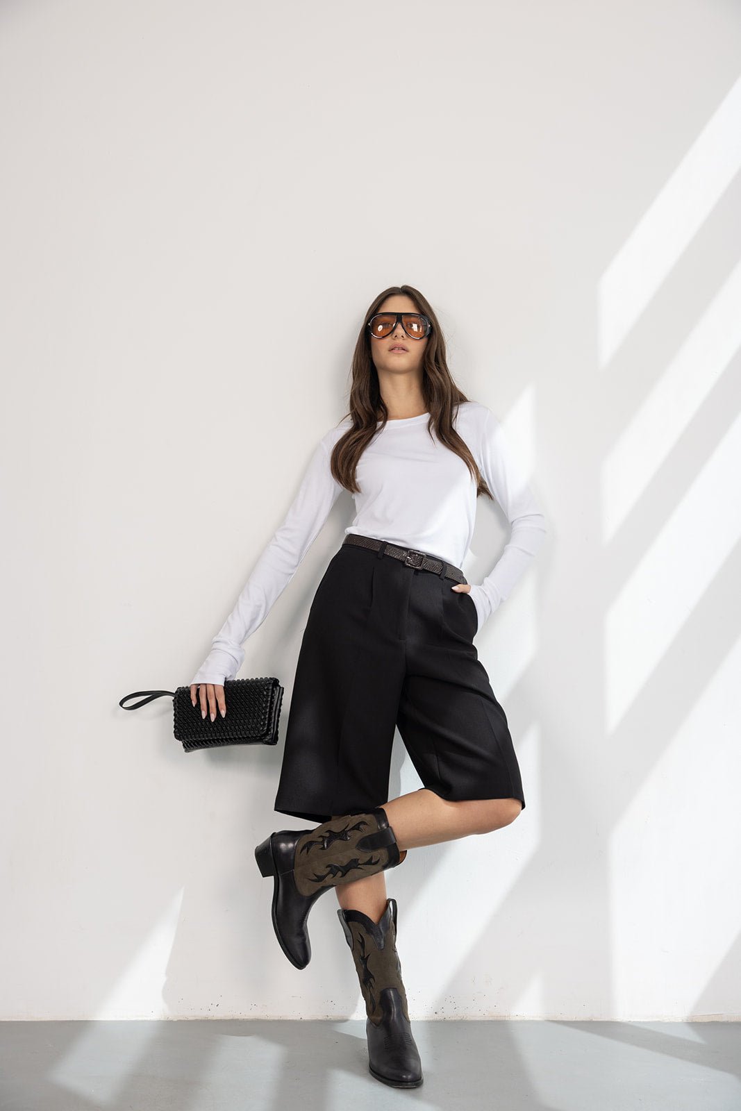 אישה עם חולצה לבנה, מכנסי ברמודה שחורים, מגפי בוקרים, תיק שחור ומשקפי שמש אופנתיים