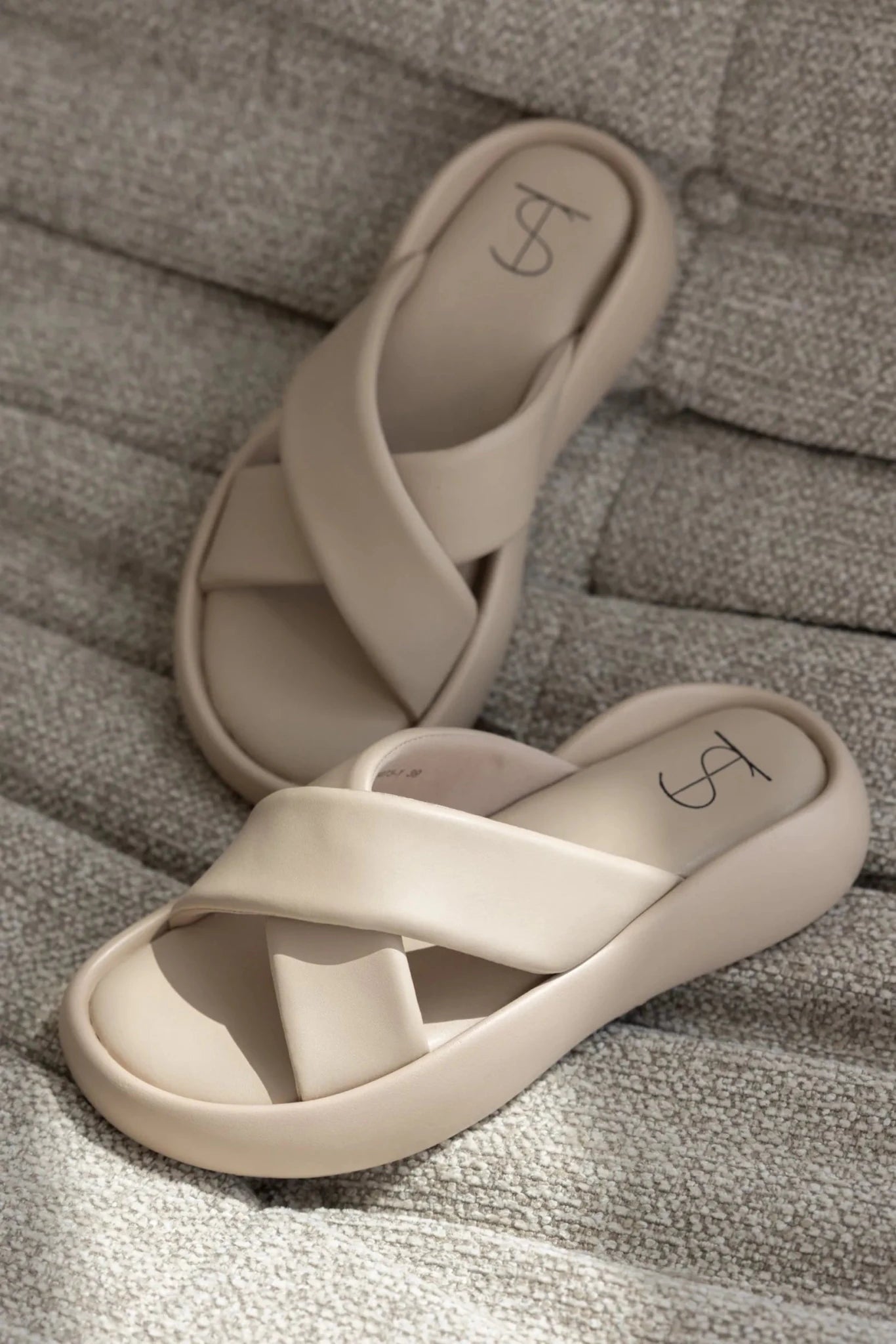 כפכפי נשים קרם עם רצועות מצטלבות, מונחים על ספה בד בהירה, אופנה טרנדית