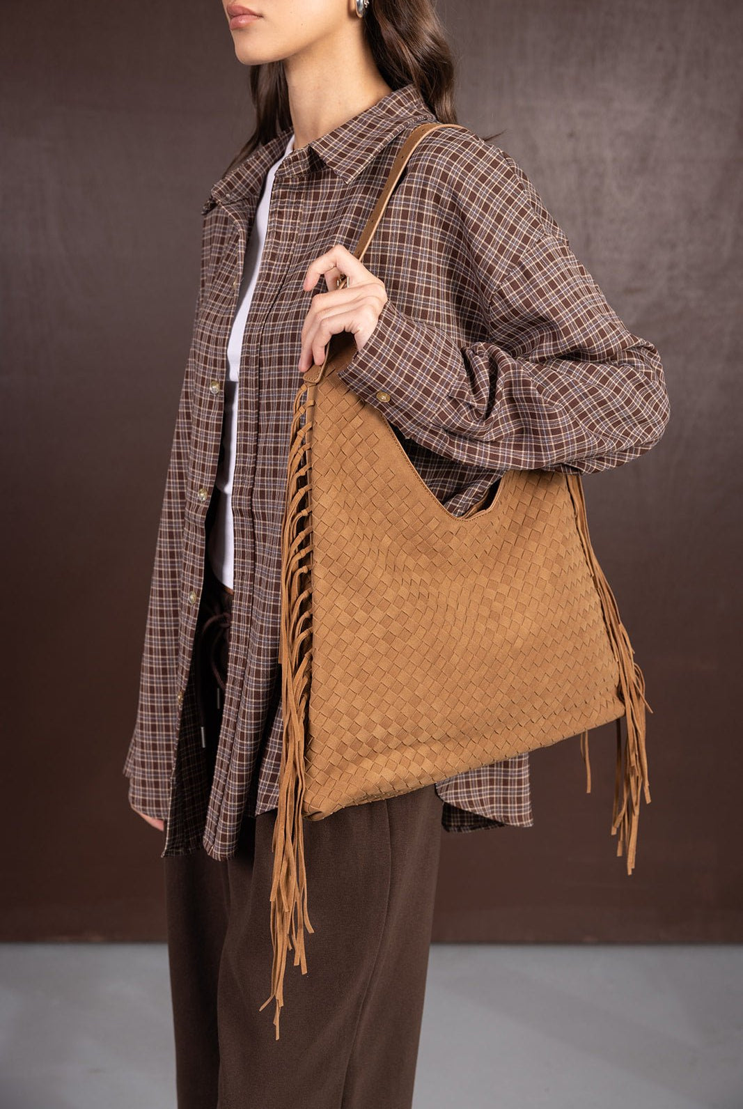 אישה לובשת חולצת משבצות חום ונושאת תיק כתף חום עם קליעה ופרנזים, אופנת נשים עדכנית