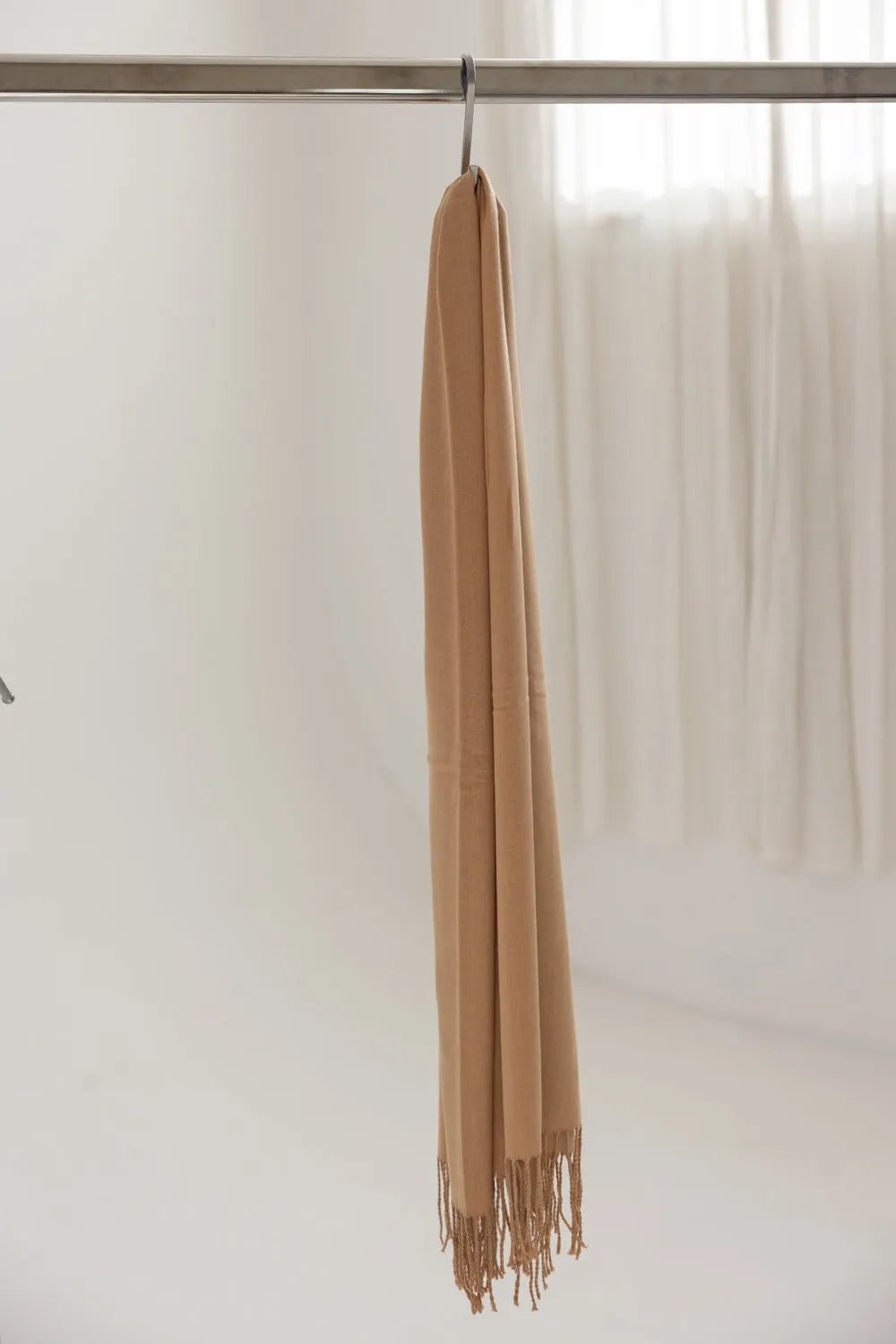 צעיף נשים בצבע קאמל, תלוי על קולב, עם קצוות פרנזים, רקע בהיר מינימליסטי