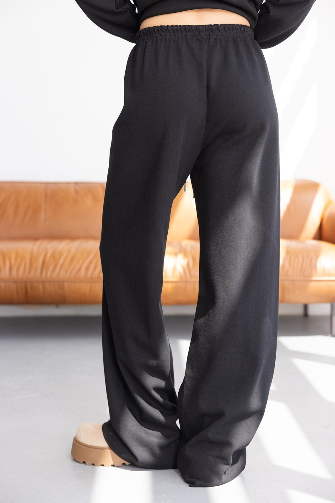 מכנסי נשים שחורים בגזרה רחבה וקומות גבוה, שיק נינוח ליד ספה עור חומה