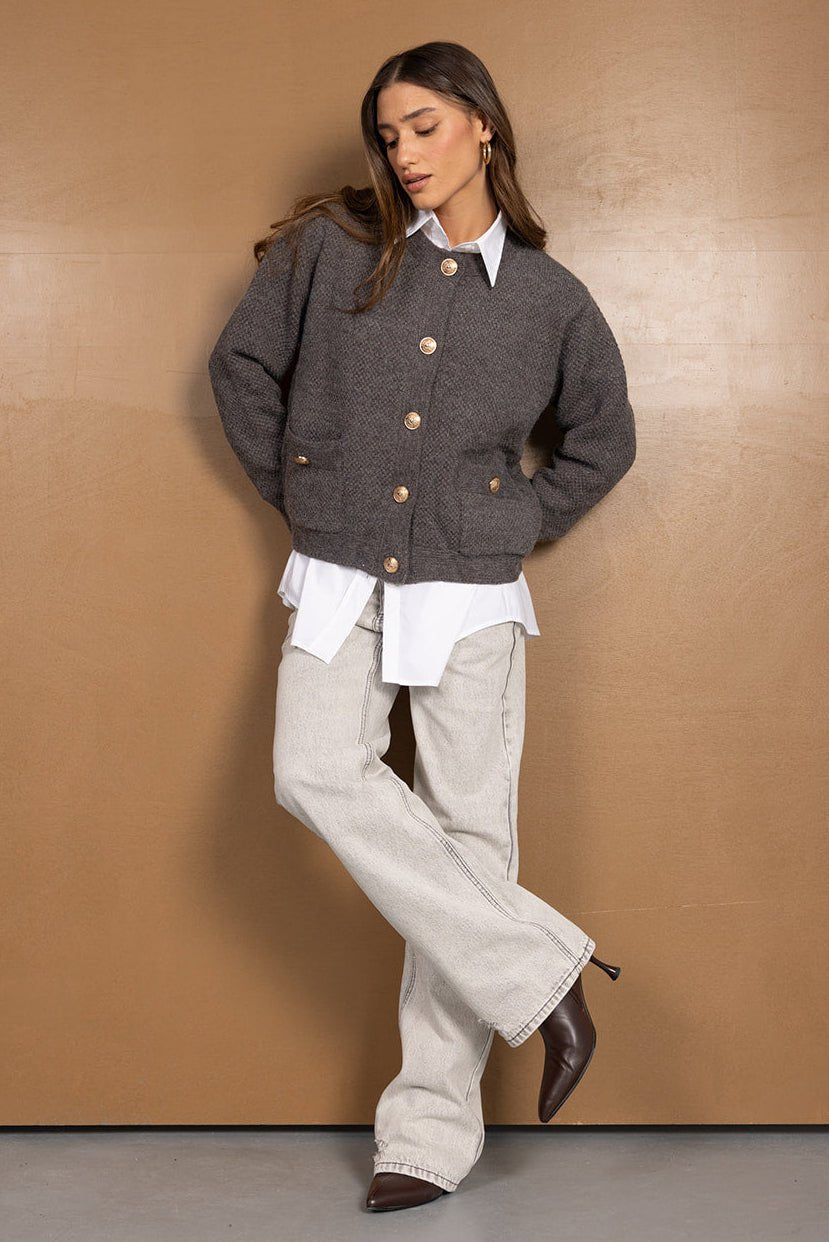 אישה דוגמנית לובשת סוודר אפור עם כפתורים, חולצה לבנה רחבה וג'ינס בהיר אופנתי.