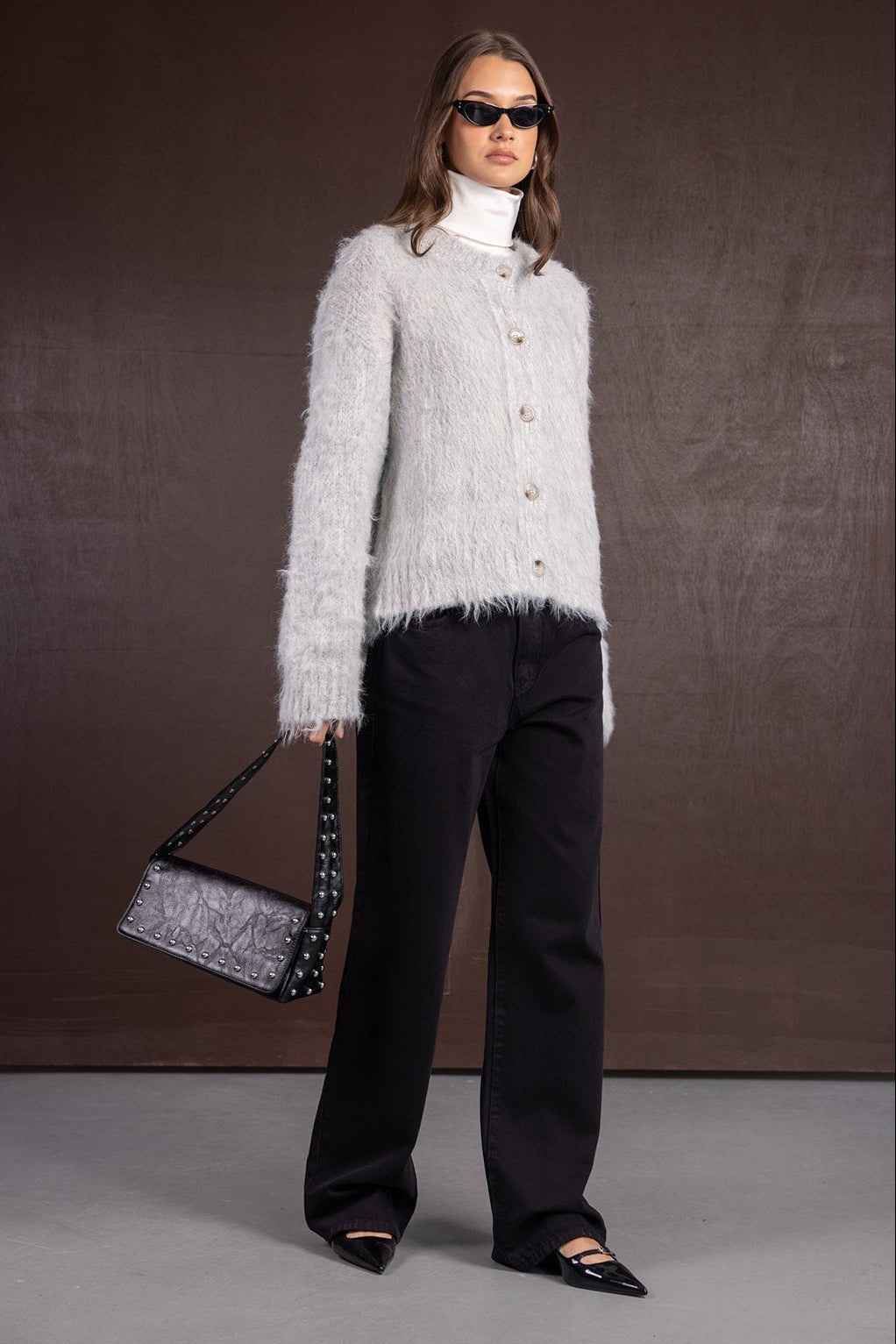 אישה עם קרדיגן פרוותי אפור, מכנסיים שחורים, גולף לבן, משקפי שמש ותיק כתף שחור, אופנת נשים חורף