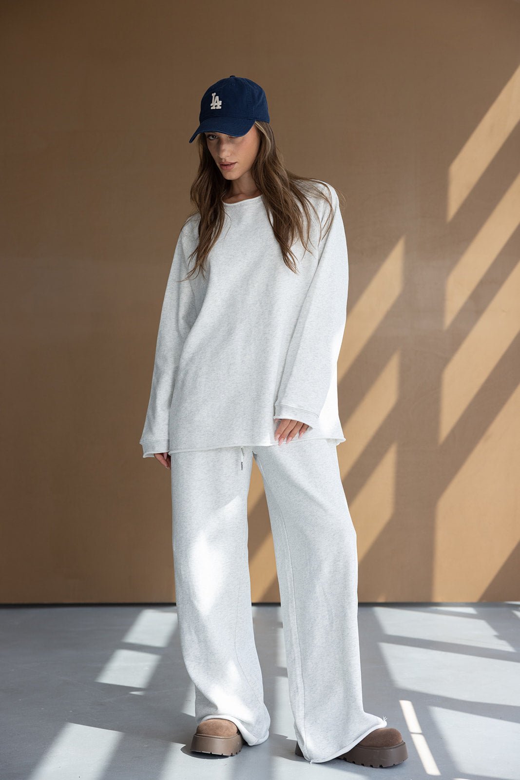 דוגמנית לובשת סט טרנינג אפור רחב, כובע מצחייה כחול ונעליים חומות, אופנה לנשים