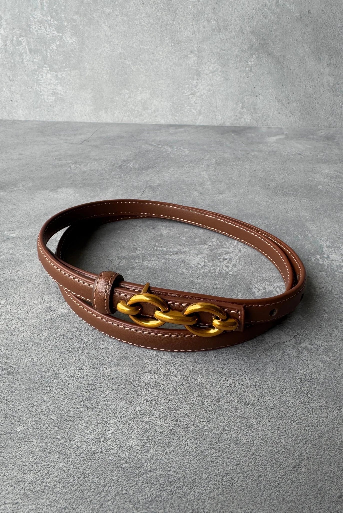 חגורת עור חומה עם אבזם שרשרת זהוב, אופנה לנשים ונערות, מונחת על משטח בטון