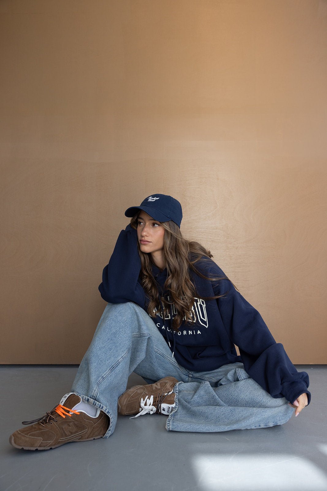 צעירה יושבת עם קפוצ׳ון כחול, כובע מצחייה, ג'ינס רחב ונעלי סניקרס חומות, אופנה טרנדית לנערות