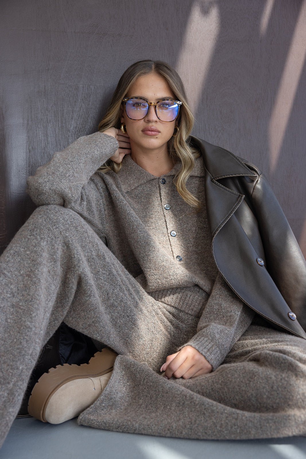 דוגמנית עם משקפי אופנה, לובשת סט טריקו חום, מעיל עור כהה ונעלי פלטפורמה, בסגנון חורפי עדכני