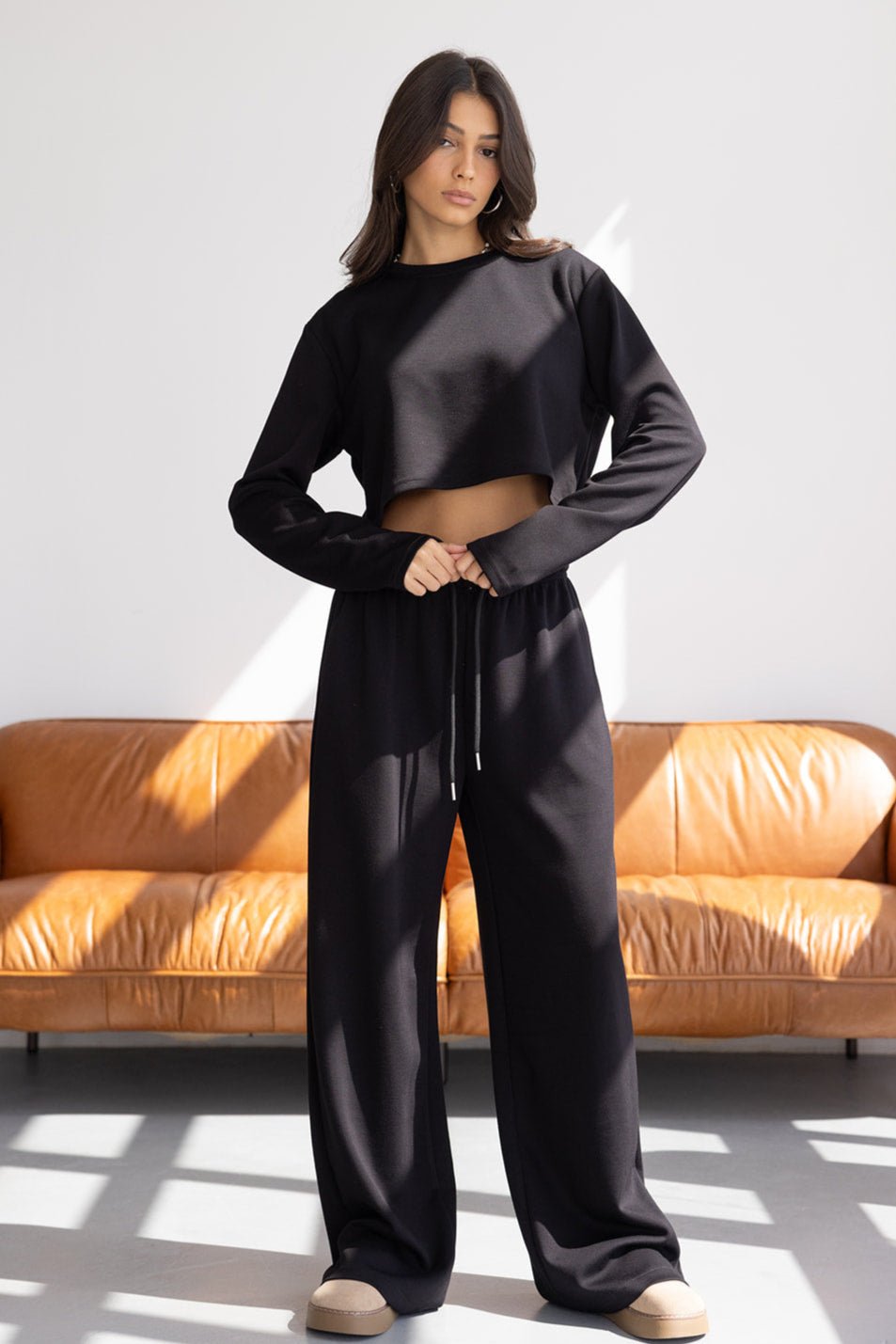 אישה צעירה לובשת סט טרנינג שחור עם חולצת קרופ ומכנסיים רחבים, עומדת בסלון מודרני