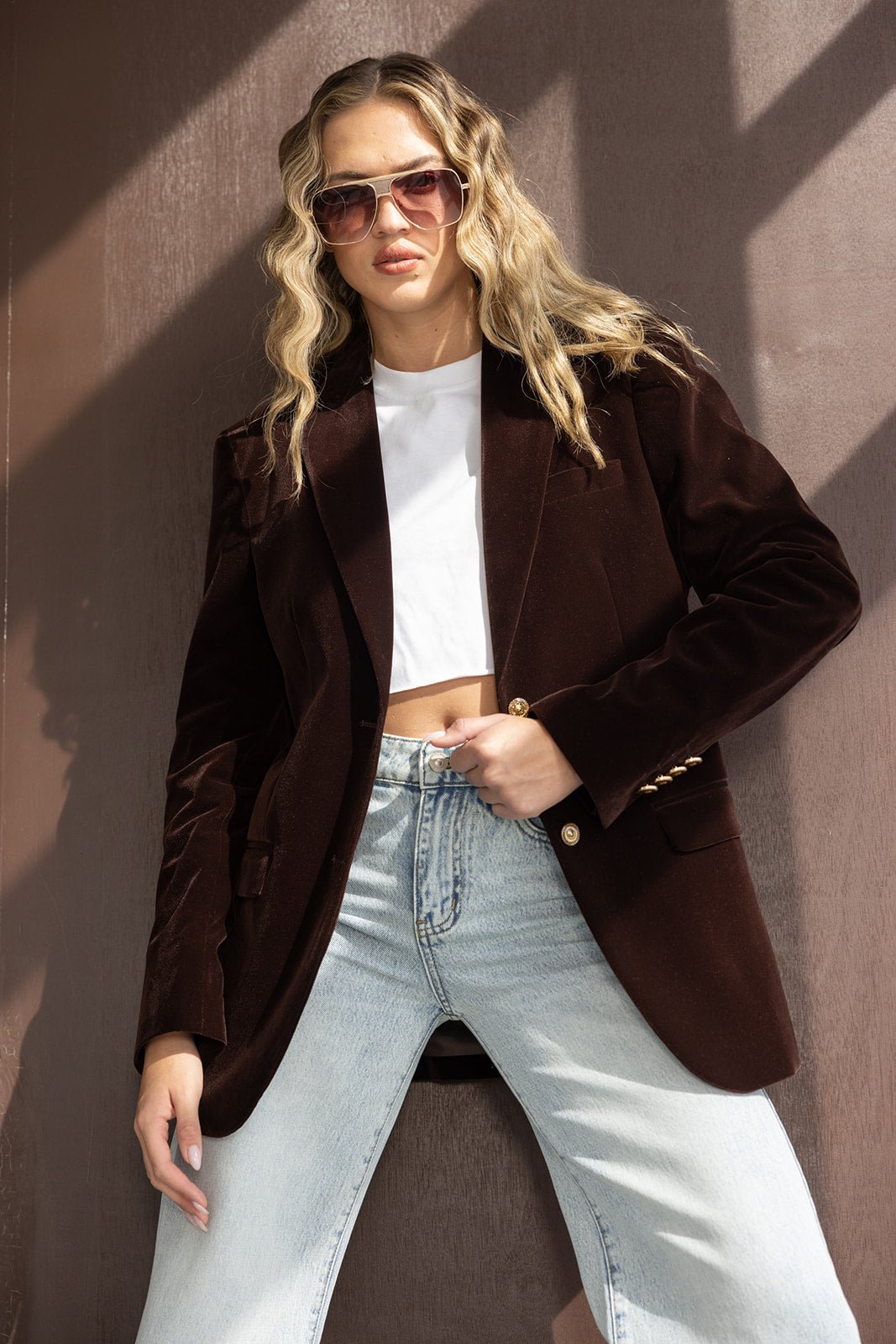 אישה עם בלייזר חום, ג'ינס בהיר, חולצה לבנה קצרה ומשקפי שמש אופנתיים, מראה רחוב טרנדי לנשים