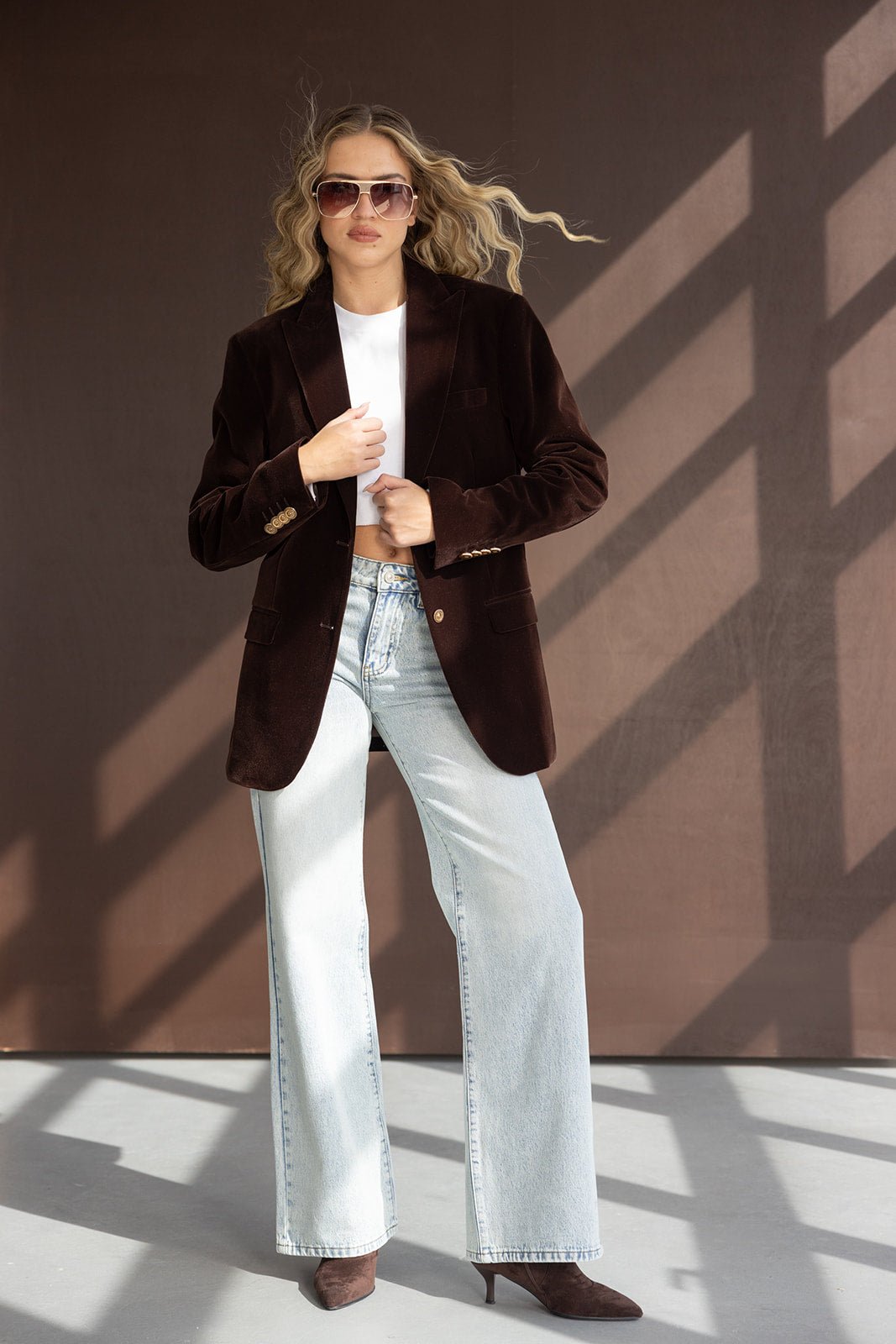 אישה עם ז'קט קטיפה חום, חולצה לבנה, ג'ינס רחב ומשקפי שמש אופנתיים