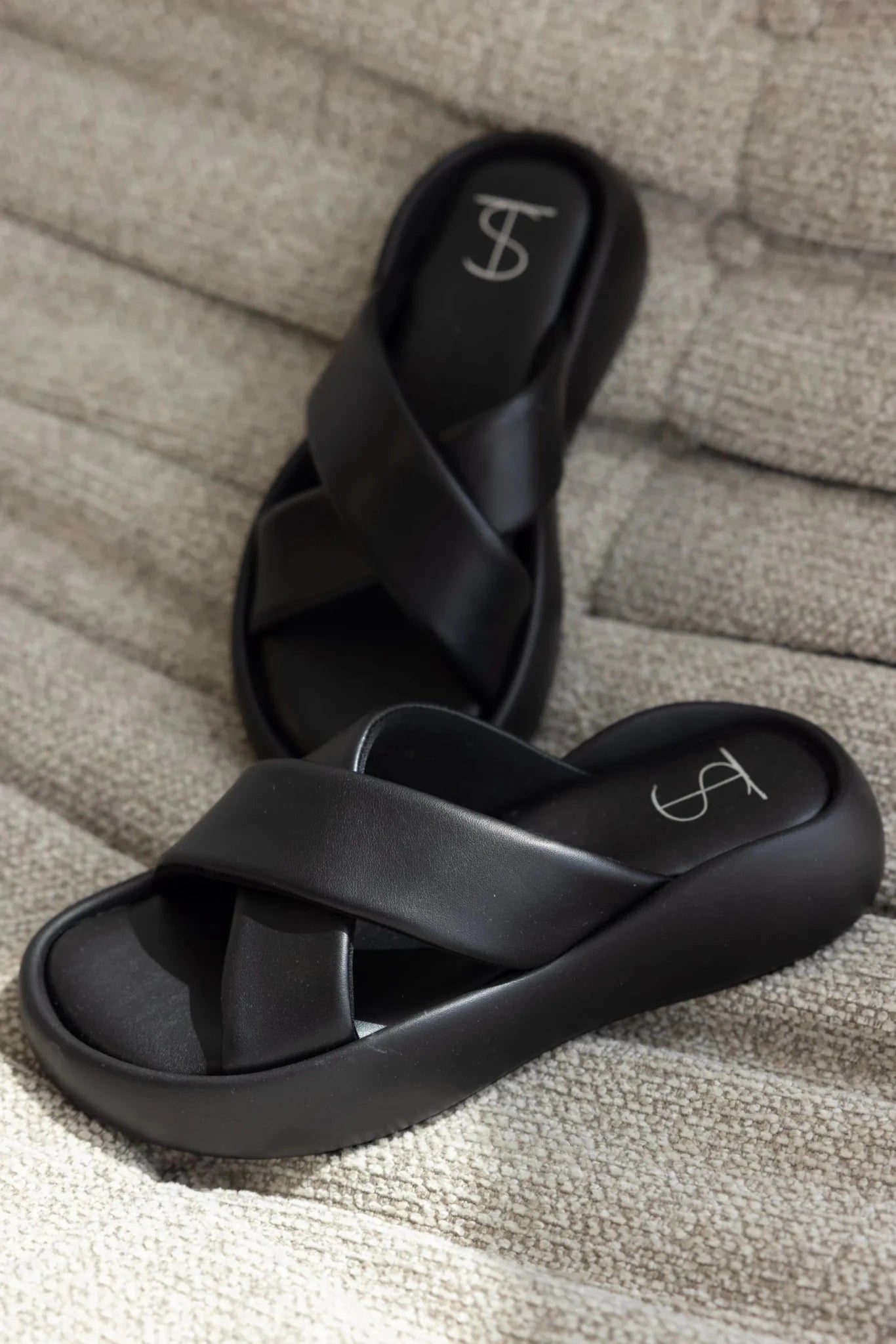 כפכפי נשים אופנתיים בצבע שחור, רצועות עבות מצטלבות, מונחים על ספה בהירה