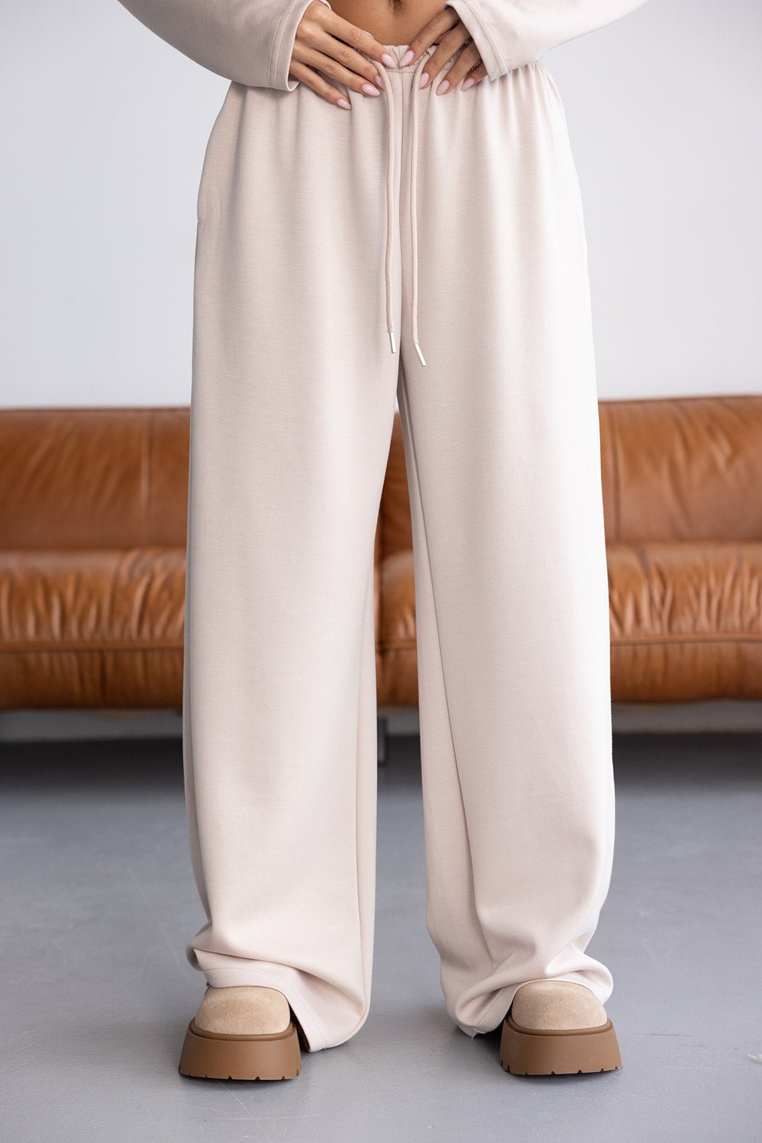 מכנסי טרנינג רחבים בצבע בז' לנשים, גומי במותן, נעלי פלטפורמה, רקע סלון חום