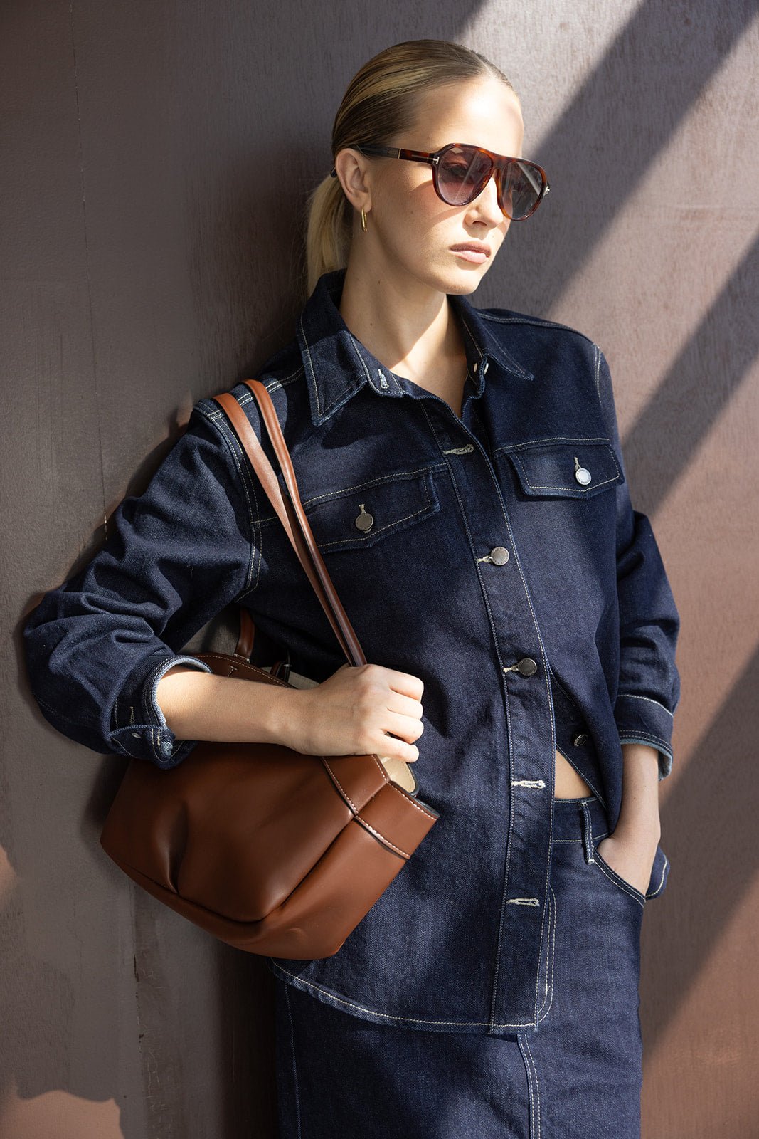 אישה עם חולצת ג'ינס כהה, חצאית תואמת, תיק חום ומשקפי שמש אופנתיים