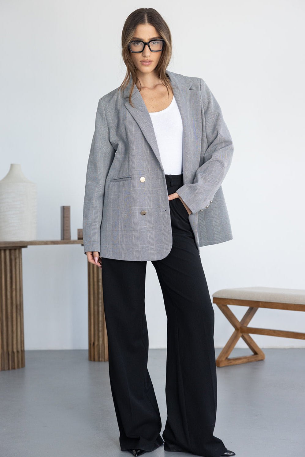 אישה עם ז'קט מחויט אפור, חולצה לבנה ומכנסיים שחורים אופנתיים בצילום סטודיו מודרני