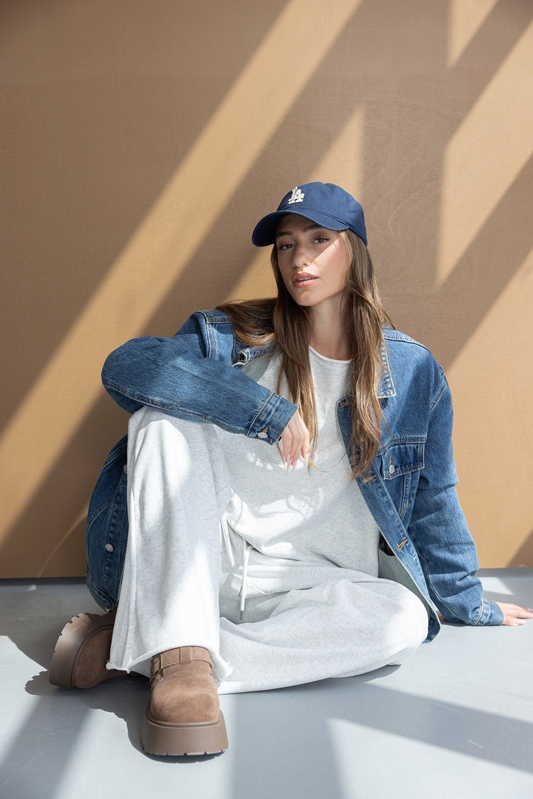 אישה יושבת על הרצפה בלבוש אופנתי, עם ג'קט ג'ינס, כובע כחול, מכנסיים לבנים ונעליים חומות