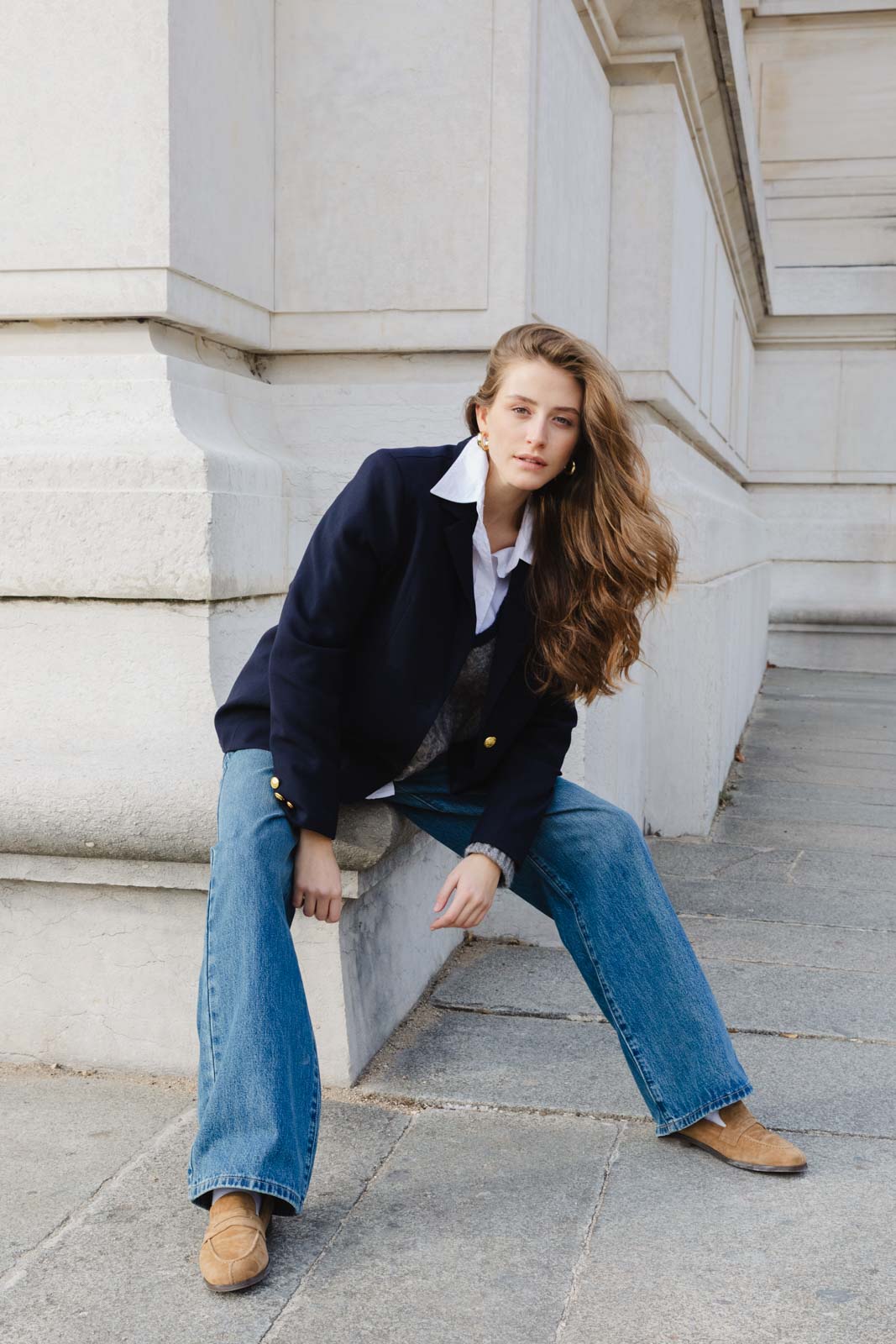 אישה צעירה באופנת רחוב עם ג'ינס, ז'קט כהה ונעליים חומות יושבת ליד קיר אבן אורבני