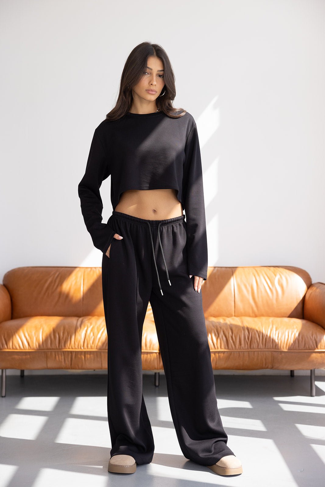 דוגמנית לובשת סט טרנינג שחור לנשים בגזרת קרופ ומכנסיים רחבים, עומדת ליד ספה חומה בסלון מואר