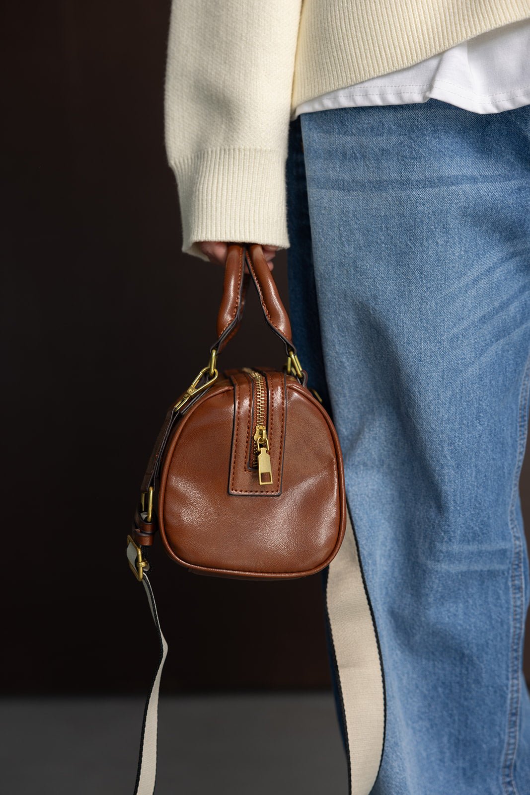 תיק עור חום אופנתי עם רוכסן זהב, ידיות קצרות ורצועה, משולב עם ג'ינס וסוודר בהיר