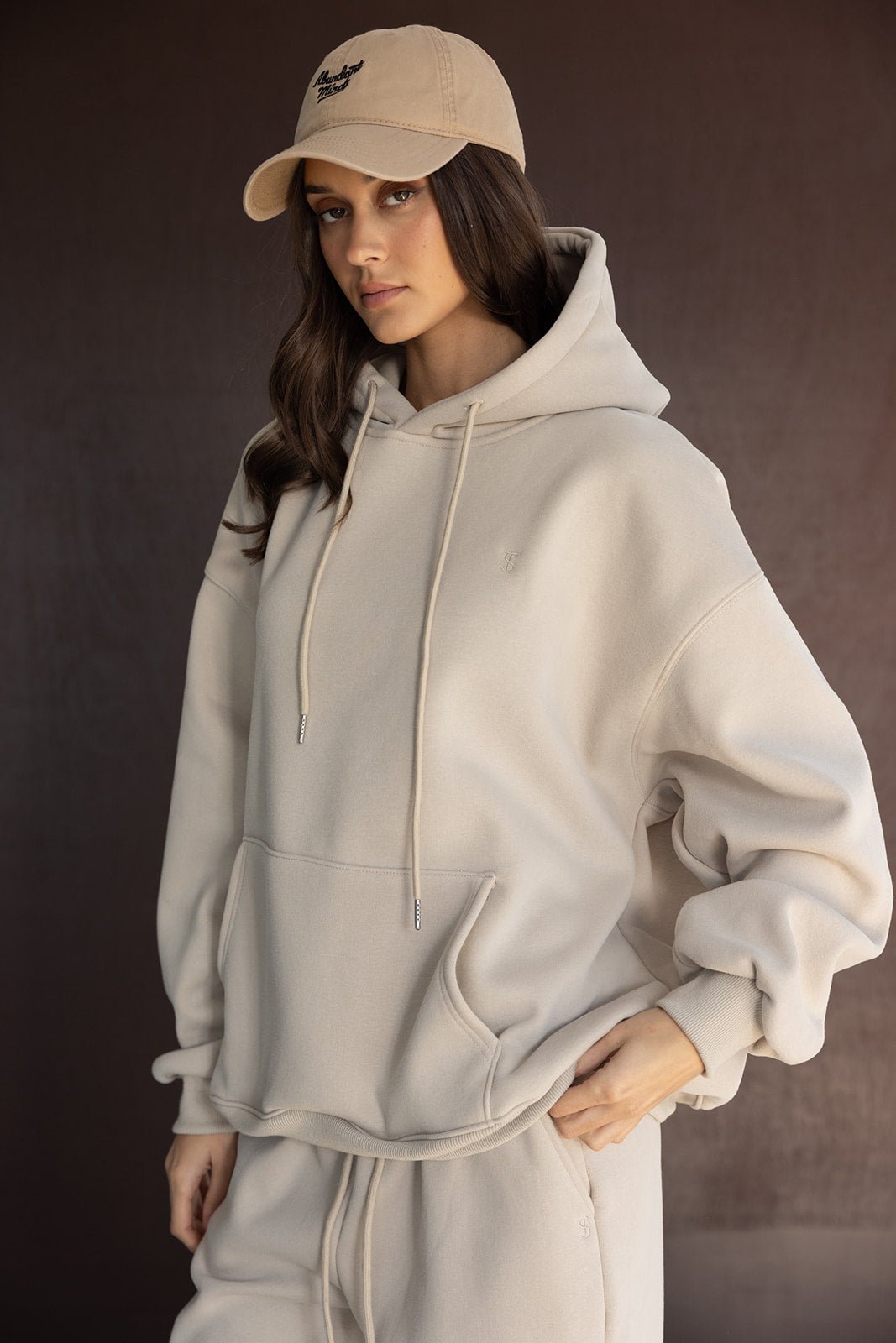 אישה לובשת סט טרנינג אופנתי בצבע בז' עם קפוצ'ון וכובע מצחייה, בגדי נשים חורף