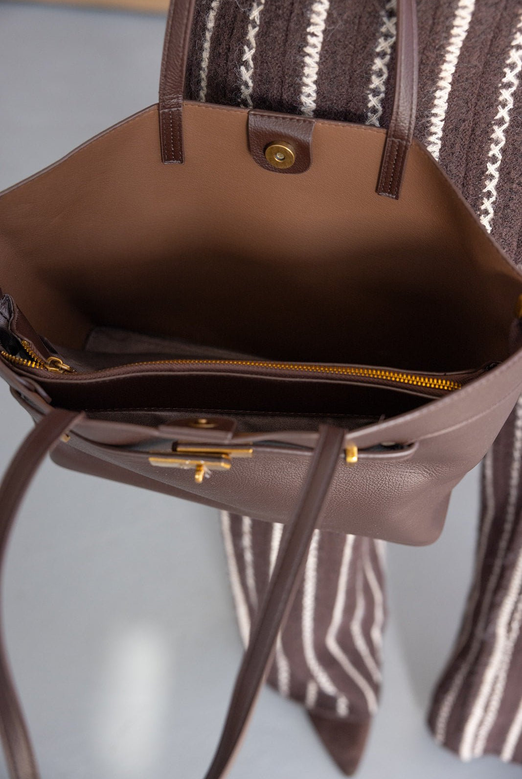 תיק עור חום אופנתי עם רוכסן וכפתור זהב, מבט על פנים התיק, על רקע בגדי נשים מפוספסים