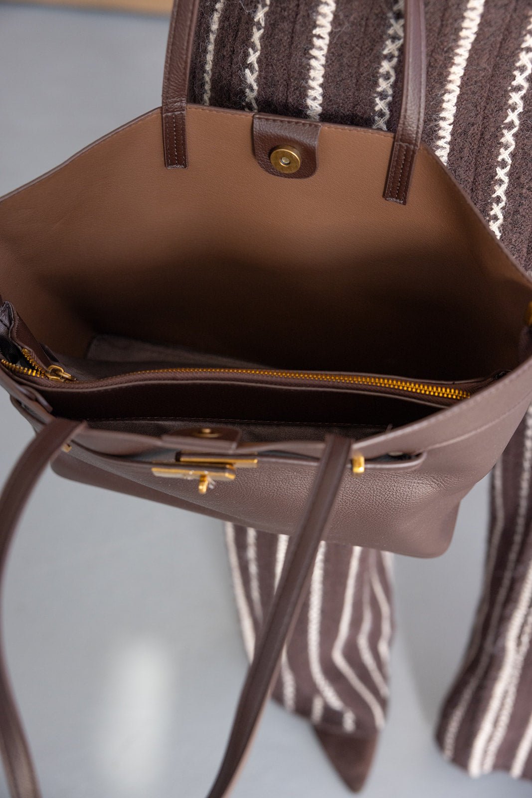 תיק עור חום אופנתי עם רוכסן וכפתור זהב, מבט על פנים התיק, על רקע בגדי נשים מפוספסים