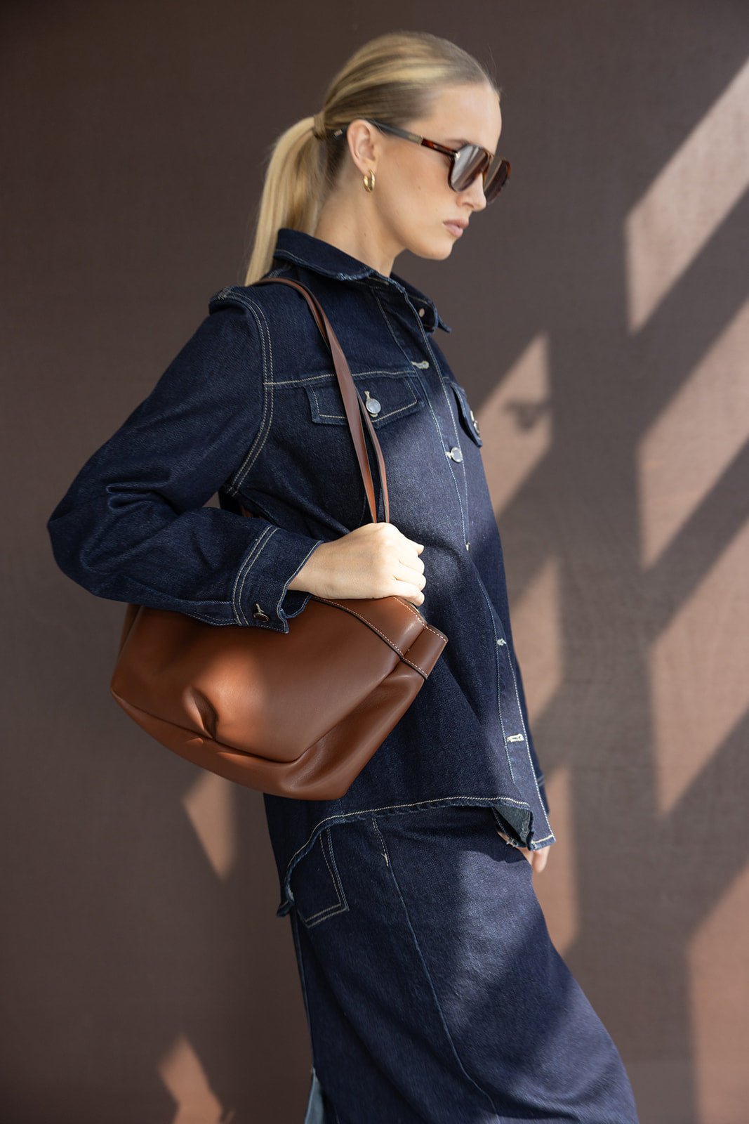 אישה לובשת ג'קט ג'ינס כהה עם תיק כתף חום אופנתי ומשקפי שמש, מראה אופנה עדכנית